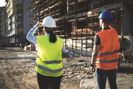 A female construction worker in a yellow hi-vis vest and white hard hat, and a male worker in an orange hi-vis vest and blue hard hat inspect a construction site.