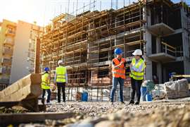 Four construction workers in hi-vis and hard hats examine documents on a construction site