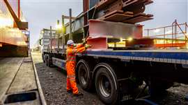 Driver Wearing Full PPE and Chaining Down a Load of Steel Structures onto a Semi Truck Flat bed with Metal Posts Fitted.