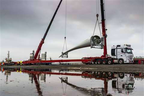 Pair of red teleboom cranes lifting a turbine blade off a red beam trailer refleted in a puddle
