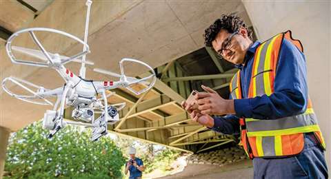 A drone pilot wearing a yellow hi-vis vest launches a drone under a concrete bridge