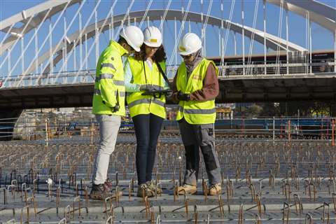 Three Ferrovial workers, one male (left) and two female, on a construction site wearing branded yellow hi-vis vests and white hard hats