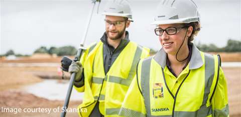 Two Skanska employees - one male and one female - wearing yellow hi-vis vests and white hard hats smile on a construction site