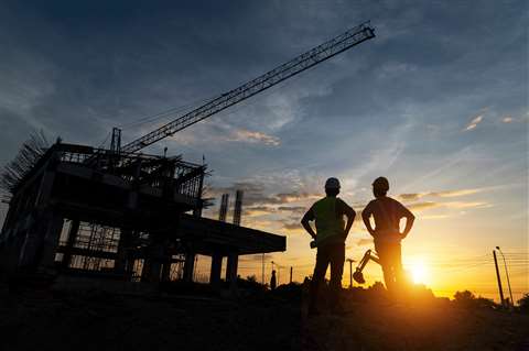 Two construction workers stand in silhouette against a construction site at sundown