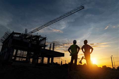 Two construction workers stand in silhouette against a construction site at sundown