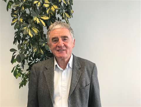 Paul Brown in a grey herringbone jacket standing in an office in front of a big green plant