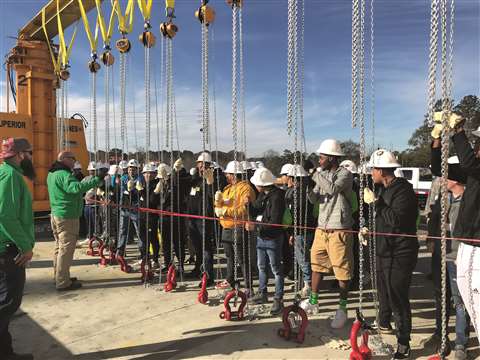 A row of multi-coloured chain hoists suspended from a beam outside in the sun surrounded by a group of trainees